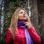 A woman in a red coat and purple scarf poses thoughtfully in a forest.